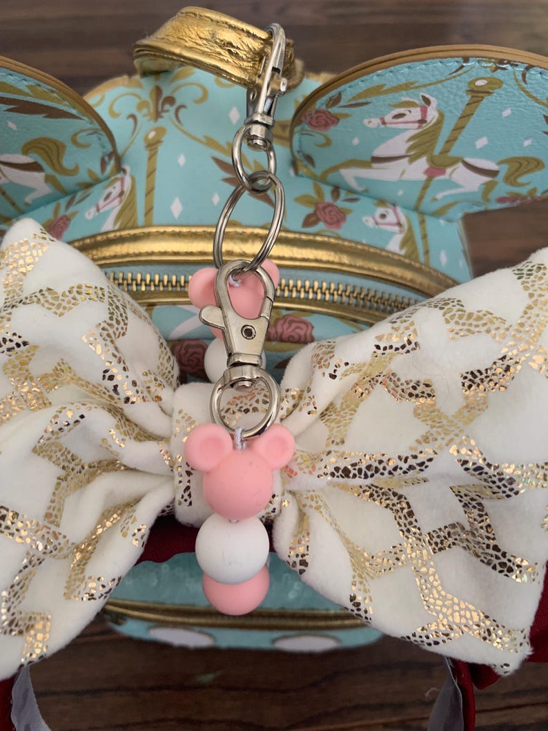 May include: A white and gold bow with a pink and white beaded charm attached to a silver key chain. The charm features a pink Mickey Mouse head.