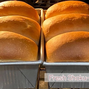 May include: Two rectangular metal loaf pans each containing three golden-brown baked bread loaves. The bread has a smooth, rounded top crust. The text "Fresh Shoku-Pan" is visible in the lower right corner of the image, set against the metallic surface of the pans.
