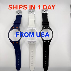 May include: Three silicone watch cases with integrated straps in navy blue, white, and black. The navy case features orange accents, while the black case has white accents. Each includes a silver-tone buckle. Text reads: SHIPS IN 1 DAY FROM USA.