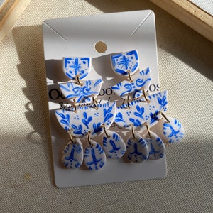 May include: A pair of blue and white ceramic inspired dangle earrings. Each earring has four  tiers with a floral design. The top tier is a small, rounded shape. The middle tier is a larger, crescent-shaped piece. The bottom tier has three teardrop-shaped pieces. The earrings are attached to gold jump rings.