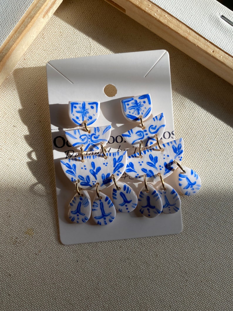 May include: A pair of blue and white ceramic inspired dangle earrings. Each earring has four  tiers with a floral design. The top tier is a small, rounded shape. The middle tier is a larger, crescent-shaped piece. The bottom tier has three teardrop-shaped pieces. The earrings are attached to gold jump rings.