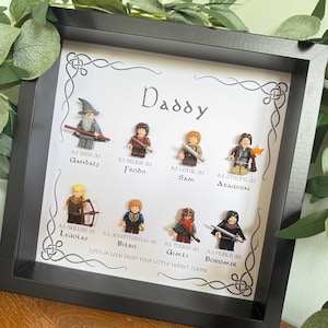 May include: A framed artwork with Lego figures of Lord of the Rings characters. The word "Daddy" is at the top, with character names and descriptions below. The frame is black, and the background is white with decorative Celtic knot borders. The text reads "Lots of love from your little hobbit, Naomi."