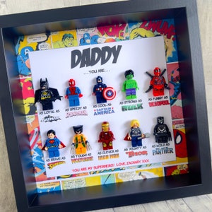 Könnte beinhalten: Ein gerahmtes Kunstwerk mit Lego-Superheldenfiguren, darunter Batman, Spider-Man und Captain America. Das Kunstwerk enthält den Text "DADDY...YOU ARE..." mit Superhelden-Vergleichen. Der Hintergrund ist im Comic-Stil gestaltet.