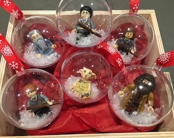 Könnte beinhalten: Sechs transparente gläserne Weihnachtskugeln mit roten und weißen Schleifenaufhängern. Jede Kugel enthält eine andere Lego-Minifigur aus der Harry-Potter-Serie. Die Minifiguren sind Harry Potter, Ron Weasley, Hermine Granger, Dobby, Severus Snape und Albus Dumbledore.