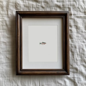 May include: A framed watercolor print of a fish. The artwork features a small, colorful fish with shades of green, pink, and brown against a white background. The frame is a dark brown color.