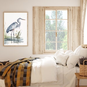 May include: A watercolor painting of a blue heron standing in a marsh. The painting is framed and hanging on a white wall above a bed with a white duvet and a yellow and brown plaid throw blanket.