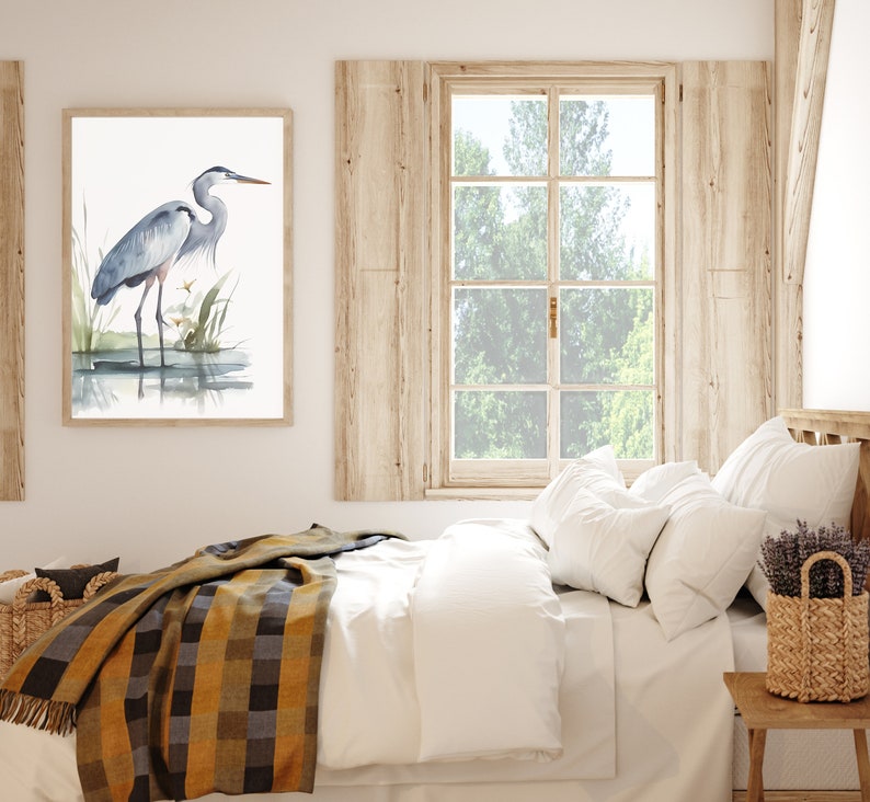 May include: A watercolor painting of a blue heron standing in a marsh. The painting is framed and hanging on a white wall above a bed with a white duvet and a yellow and brown plaid throw blanket.