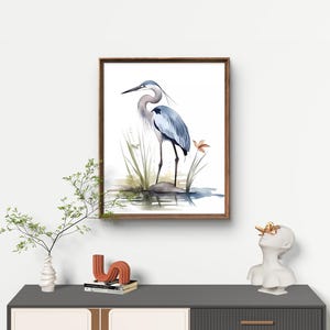 May include: A framed watercolor painting of a blue heron standing on a rock in a pond. The heron is facing to the left and has its wings folded. The painting is set against a white background.