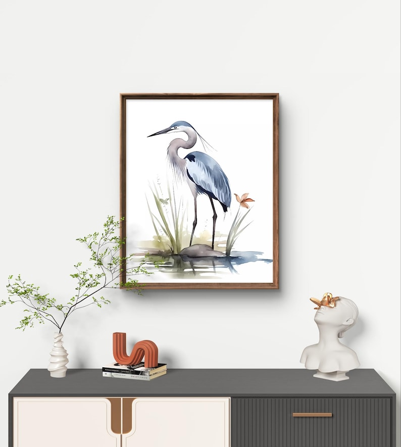 May include: A framed watercolor painting of a blue heron standing on a rock in a pond. The heron is facing to the left and has its wings folded. The painting is set against a white background.