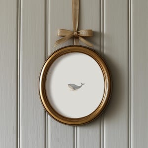 May include: A round, gold-colored picture frame with a watercolor illustration of a whale. The frame is suspended by a gold ribbon tied in a bow. The background is a light gray, vertical-paneled wall.