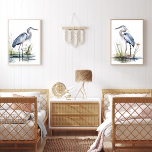 May include: Two watercolor paintings of blue herons standing in a marsh setting, framed in natural wood, hanging on a white wall above two wicker twin beds with white bedding and a woven rug on the floor.