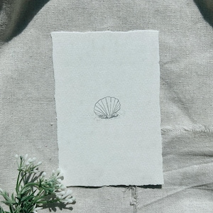 May include: A minimalist black and white illustration of a seashell on a textured, off-white paper. The artwork features a detailed outline of the shell, resting on a small patch of sand, against a neutral background.