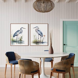 May include: Two framed watercolor paintings of blue herons standing in a marsh setting. The paintings are hanging on a white wall above a white round table with four chairs. The chairs are made of wood and have woven seats.
