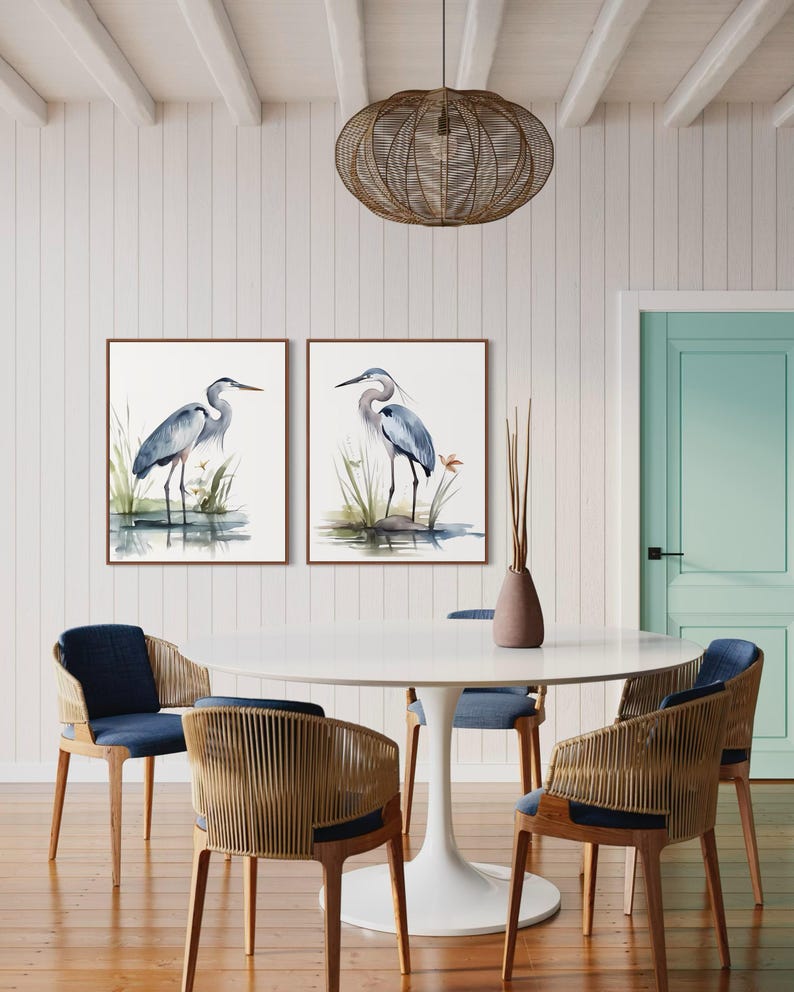 May include: Two framed watercolor paintings of blue herons standing in a marsh setting. The paintings are hanging on a white wall above a white round table with four chairs. The chairs are made of wood and have woven seats.