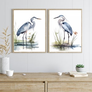 May include: Two watercolor paintings of blue herons standing in a marsh setting. The herons are depicted in a light blue and gray color scheme with a light brown background. The paintings are framed in a light brown wood frame.