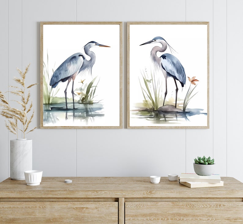 May include: Two watercolor paintings of blue herons standing in a marsh setting. The herons are depicted in a light blue and gray color scheme with a light brown background. The paintings are framed in a light brown wood frame.