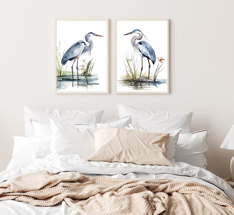 May include: Two watercolor paintings of blue herons standing in a marsh setting. The herons are depicted in a light blue and gray color scheme with a white background. The paintings are framed in a simple white frame.