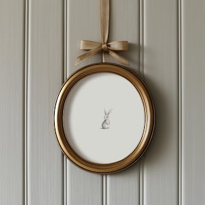 May include: A round, gold-colored picture frame with a watercolor illustration of a small bunny. The frame is suspended by a gold ribbon tied in a bow. The background is a light gray, vertical-paneled wall.