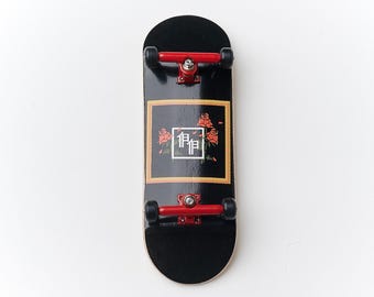 Puede incluir: Fingerboard negro con trucks rojos y un gráfico de un marco dorado con rosas rojas y el texto "1919" en su interior.