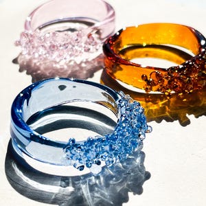 May include: Three colorful glass rings. One is pink with a textured design, another is amber with a similar design, and the third is blue with a textured design. The rings are on a white surface.