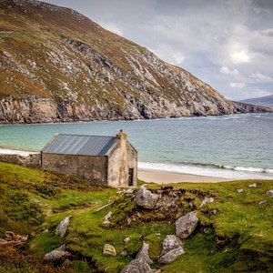 May include: A small, gray stone building with a metal roof sits on a grassy hillside overlooking a sandy beach and a body of water. The building has a single door and a window. The beach is surrounded by green hills.