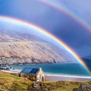 May include: A small, white cottage with a blue roof sits on a grassy hillside overlooking a sandy beach and a turquoise ocean. A double rainbow arches over the scene, creating a picturesque landscape.