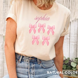 May include: A light beige t-shirt with the name "sophia" printed in pink. The shirt features six pink bows arranged in two rows of three.