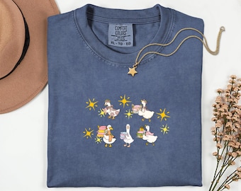 May include: A blue Comfort Colors t-shirt with a whimsical design of geese carrying books and pastries, accented with yellow starbursts. A gold star pendant necklace rests on the shirt. The shirt is size XL-TG-EG.