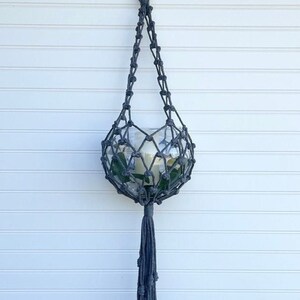 May include: A gray macrame plant hanger with a glass globe and a long tassel. The hanger is hanging from a white wall.