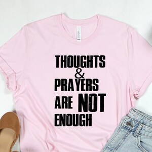 May include: Pink t-shirt with black text that reads "Thoughts & Prayers Are Not Enough".