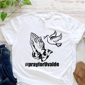 May include: White t-shirt with a black graphic of praying hands and a dove with the text #prayforUvalde.