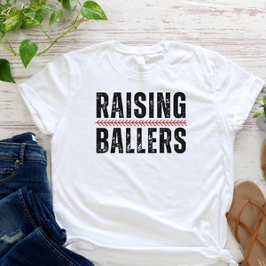 May include: White t-shirt with the text "Raising Ballers" in black with a red baseball stitch design.