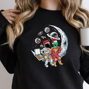 May include: Black sweatshirt with a graphic of an astronaut and an alien wearing Santa hats sitting on a crescent moon with stars in the background.