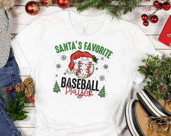 May include: White t-shirt with a green and red graphic of a baseball with a Santa hat on it. The text "Santa's Favorite Baseball Player" is printed on the shirt.