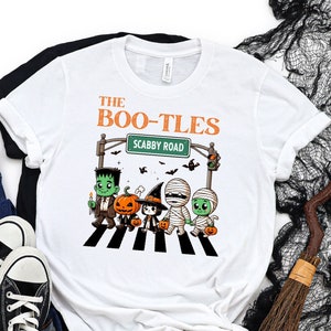 May include: White t-shirt with a graphic of a crosswalk with five cartoon characters dressed as Halloween monsters. The text "The Boo-Tles" and "Scabby Road" are above the crosswalk.