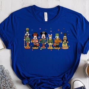 May include: Royal blue t-shirt featuring a colorful graphic of Disney characters dressed as nutcrackers. The characters include Mickey Mouse, Minnie Mouse, Donald Duck, and Goofy, each in a festive uniform. The design is set against a backdrop of small, colorful ornaments.