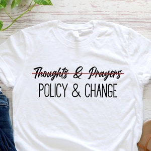 May include: White t-shirt with the text "Thoughts & Prayers" crossed out with a red line and the text "POLICY & CHANGE" below it. The shirt is laid flat on a white wooden surface with a pair of blue jeans and sandals.