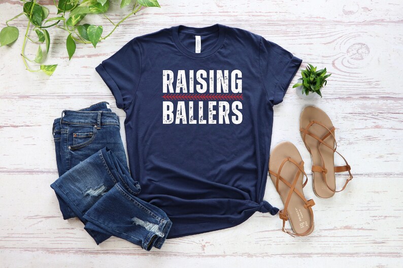 May include: A navy blue t-shirt with the text "Raising Ballers" in white with a red baseball stitch graphic.