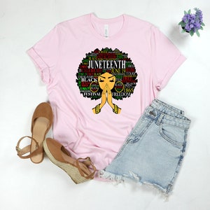 May include: Pink t-shirt with a black and green graphic of a woman's face with an afro hairstyle. The graphic is made up of words related to Juneteenth, including "Juneteenth", "Freedom", "Black", and "1865".