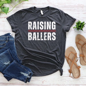 May include: A dark gray t-shirt with the text "Raising Ballers" in white with a red baseball stitch graphic above the text.