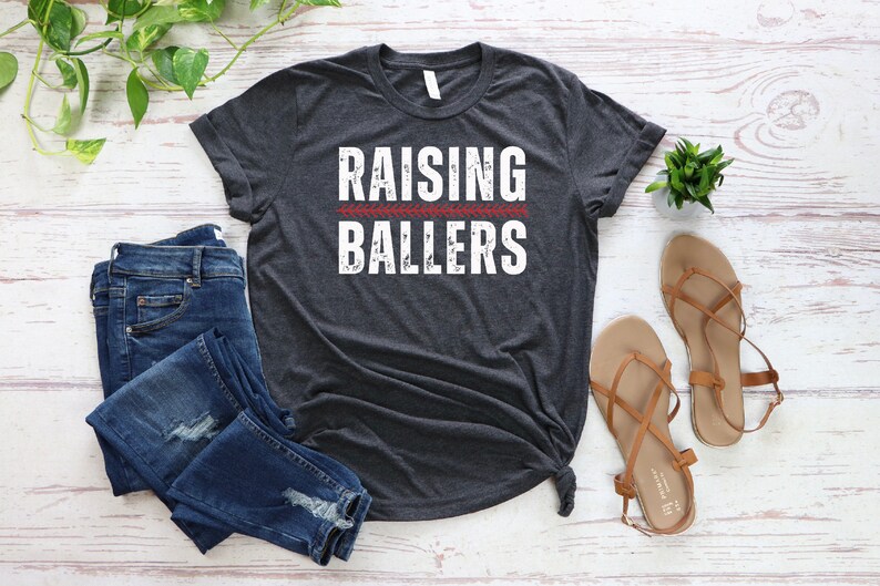 May include: A dark gray t-shirt with the text "Raising Ballers" in white with a red baseball stitch graphic above the text.