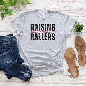 May include: A gray t-shirt with the text "Raising Ballers" in black with a red baseball stitch graphic.
