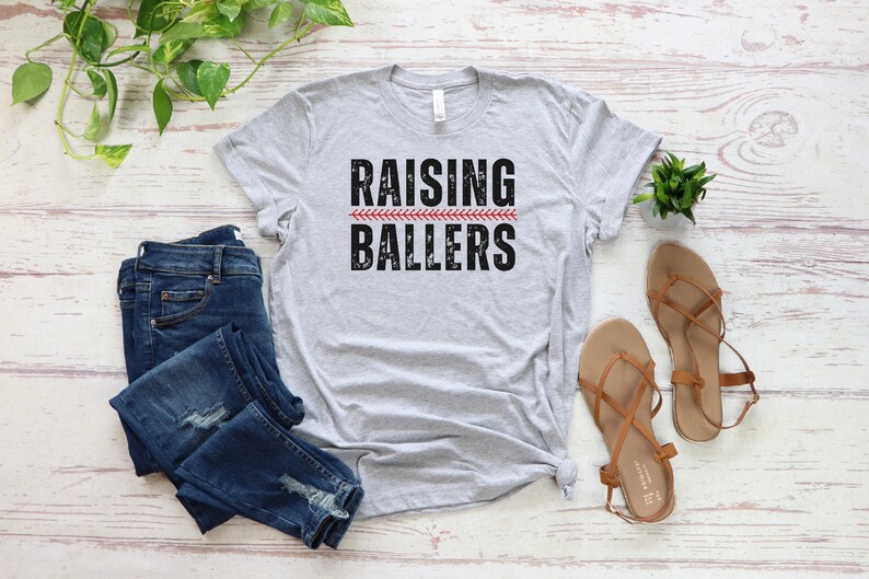 May include: A gray t-shirt with the text "Raising Ballers" in black with a red baseball stitch graphic.