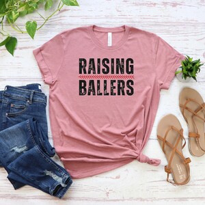 May include: A dusty rose t-shirt with the text "RAISING BALLERS" in black with a red baseball stitch graphic.