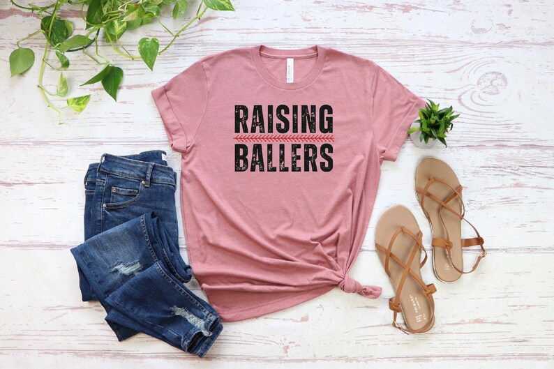 May include: A dusty rose t-shirt with the text "RAISING BALLERS" in black with a red baseball stitch graphic.