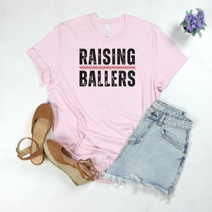 May include: Pink t-shirt with the text "RAISING BALLERS" in black with a red baseball stitch graphic.