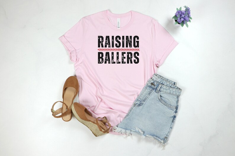 May include: Pink t-shirt with the text "RAISING BALLERS" in black with a red baseball stitch graphic.