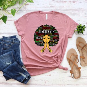 May include: A pink t-shirt with a graphic design of a black woman with an afro. The design features the word "Juneteenth" and other related words in green, red, and black. The woman is depicted with her hands clasped in prayer.
