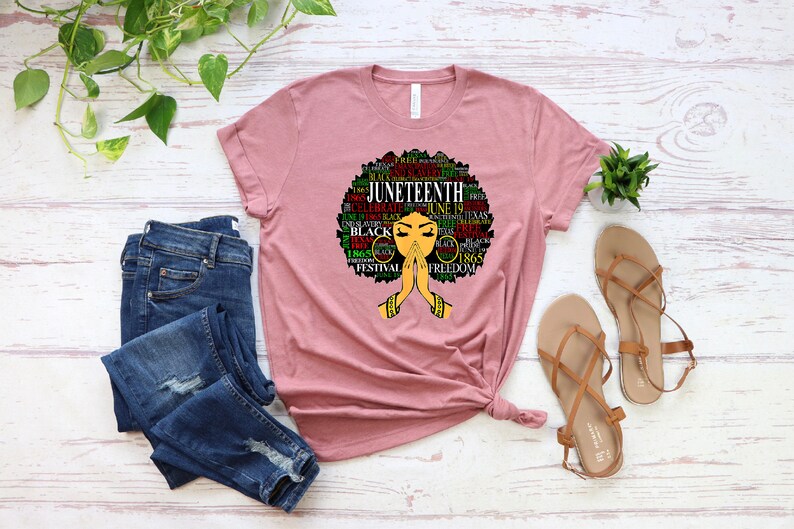 May include: A pink t-shirt with a graphic design of a black woman with an afro. The design features the word "Juneteenth" and other related words in green, red, and black. The woman is depicted with her hands clasped in prayer.