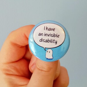 May include: A round, light blue button with the text "I have an invisible disability" in a white speech bubble. A small white ghost illustration is below the text. The button is held by a person.